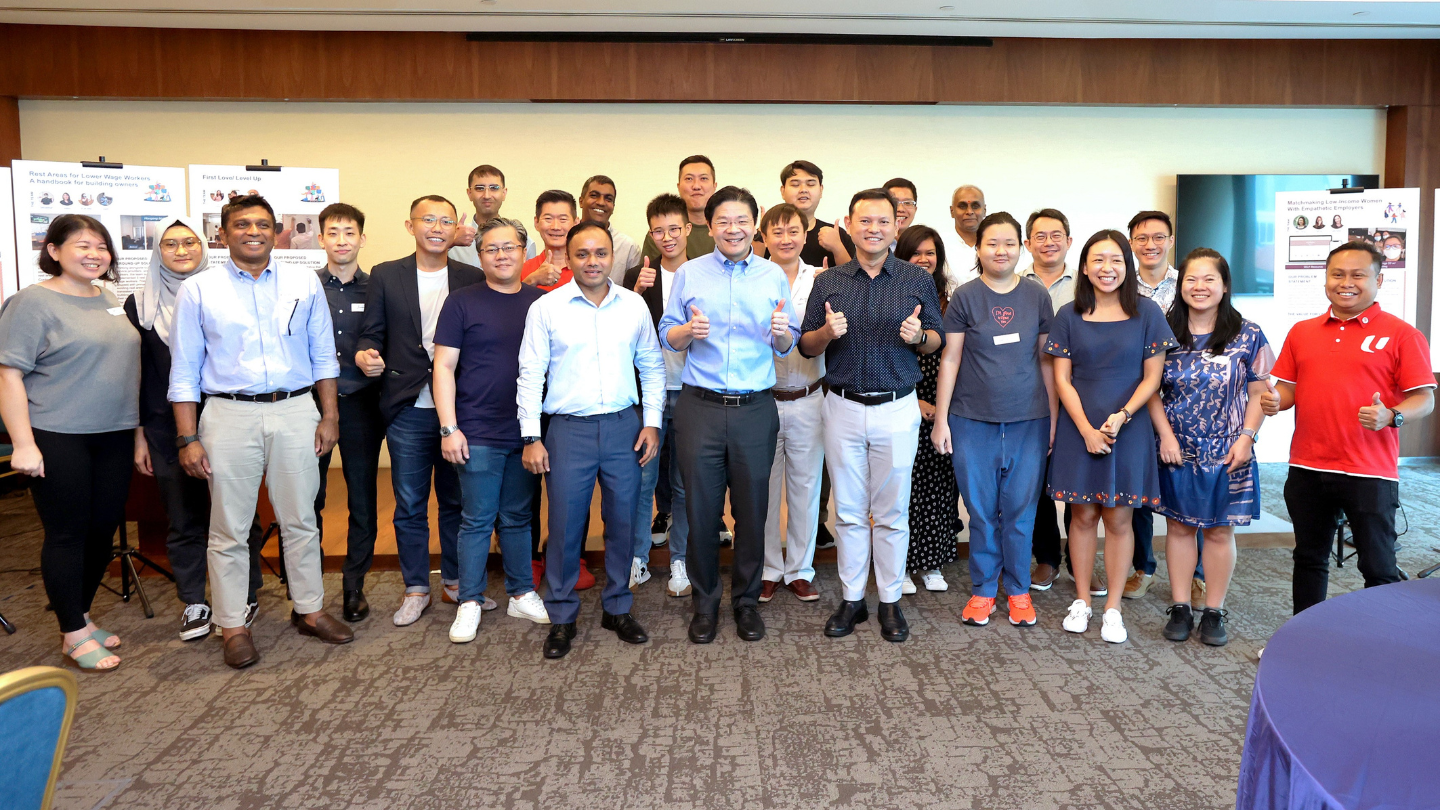 Group of diverse people posing for a photo with thumbs-up gesture at an indoor event.